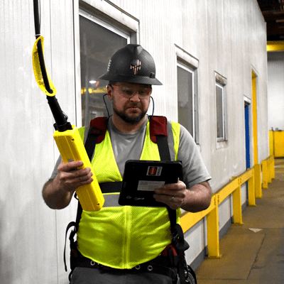 Man with black facial hair wearing safety glasses, a black hard hat, and safety green hi-vis vest over a grey t-shirt performing an inspection with an iPad for CraneWerks in Morristown, IN