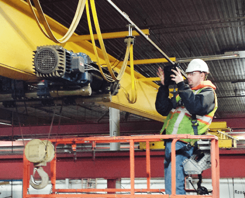 CraneWerks employee in a black hoodie, hi-vis safety green safety vest, and white hard hat performing a hoist inspection in an orange lift