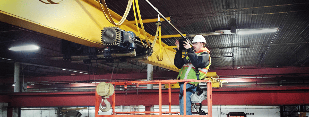 CraneWerks employee in a black hoodie, hi-vis safety green safety vest, and white hard hat performing a hoist inspection in an orange lift