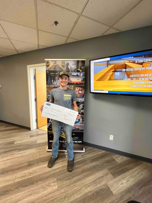 Gavin Shaw dressed in a hat, grey t-shirt, and jeans standing in the CraneWerks lobby with his oversized scholarship check from the Roger Graham Scholarship fund.