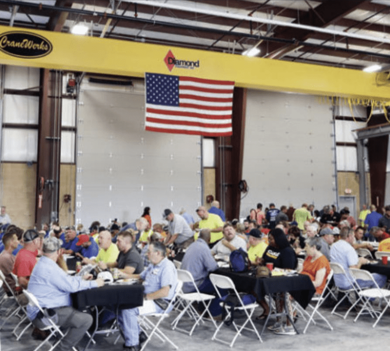 Lunch being served to a large group of people sitting under an American flag and CraneWerks overhead bridge crane at the new Diamond Equipment location.